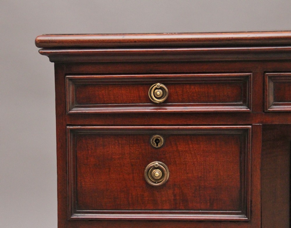 An early 19th-century mahogany kneehole desk with a dark leather writing top, multiple side drawers with circular brass ring handles, and a central recessed cupboard door. View XI