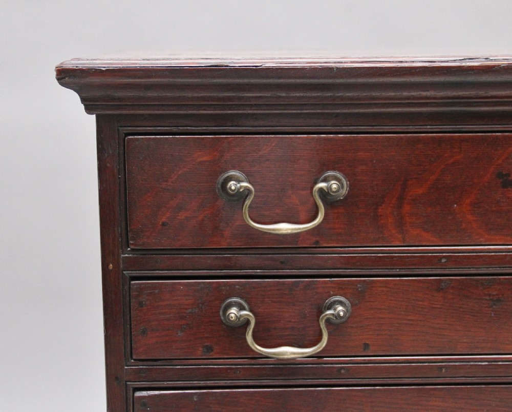 An 18th-century Georgian dark oak chest of drawers featuring five horizontal graduated drawers with brass swan-neck handles, brass escutcheons, and bracket feet. View VI