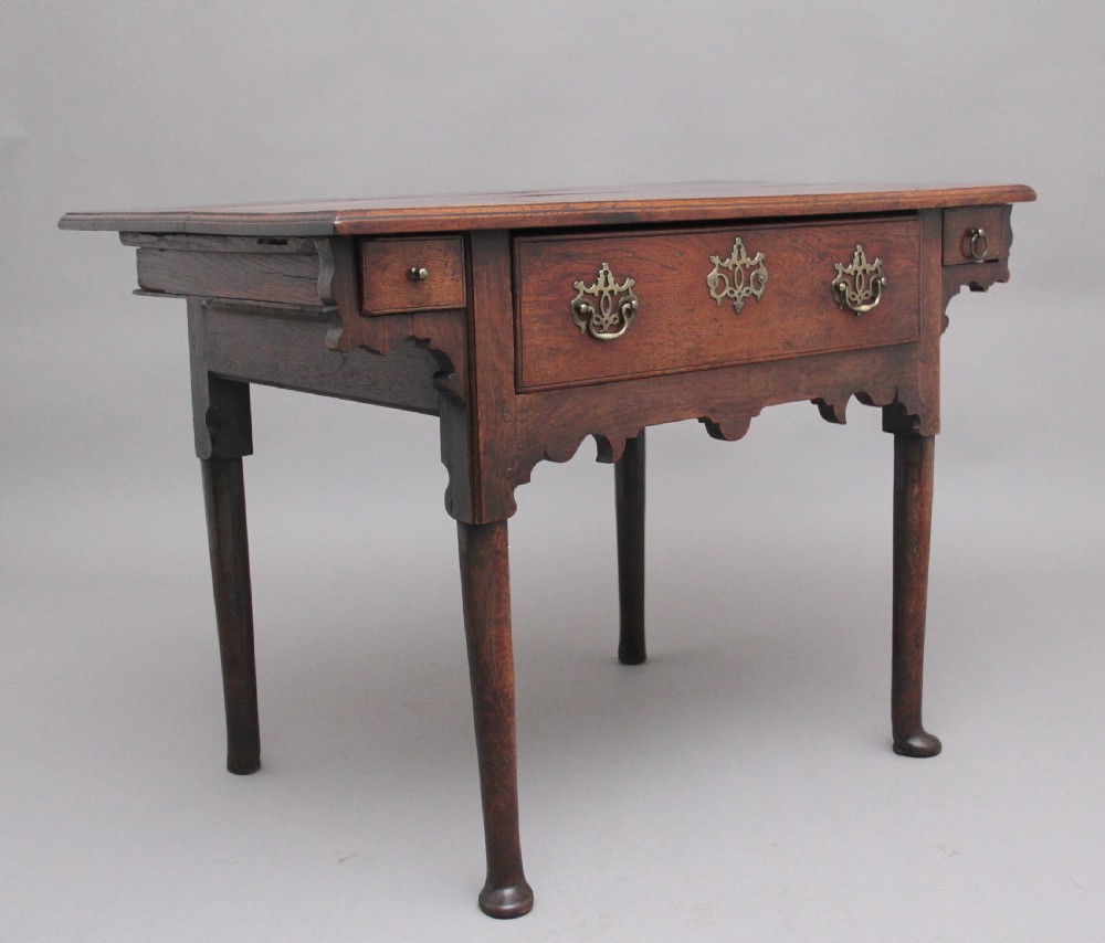 An 18th-century dark oak side table featuring a central drawer, small side drawers, pierced brass handles, a shaped apron, and turned legs ending in pad feet. View V