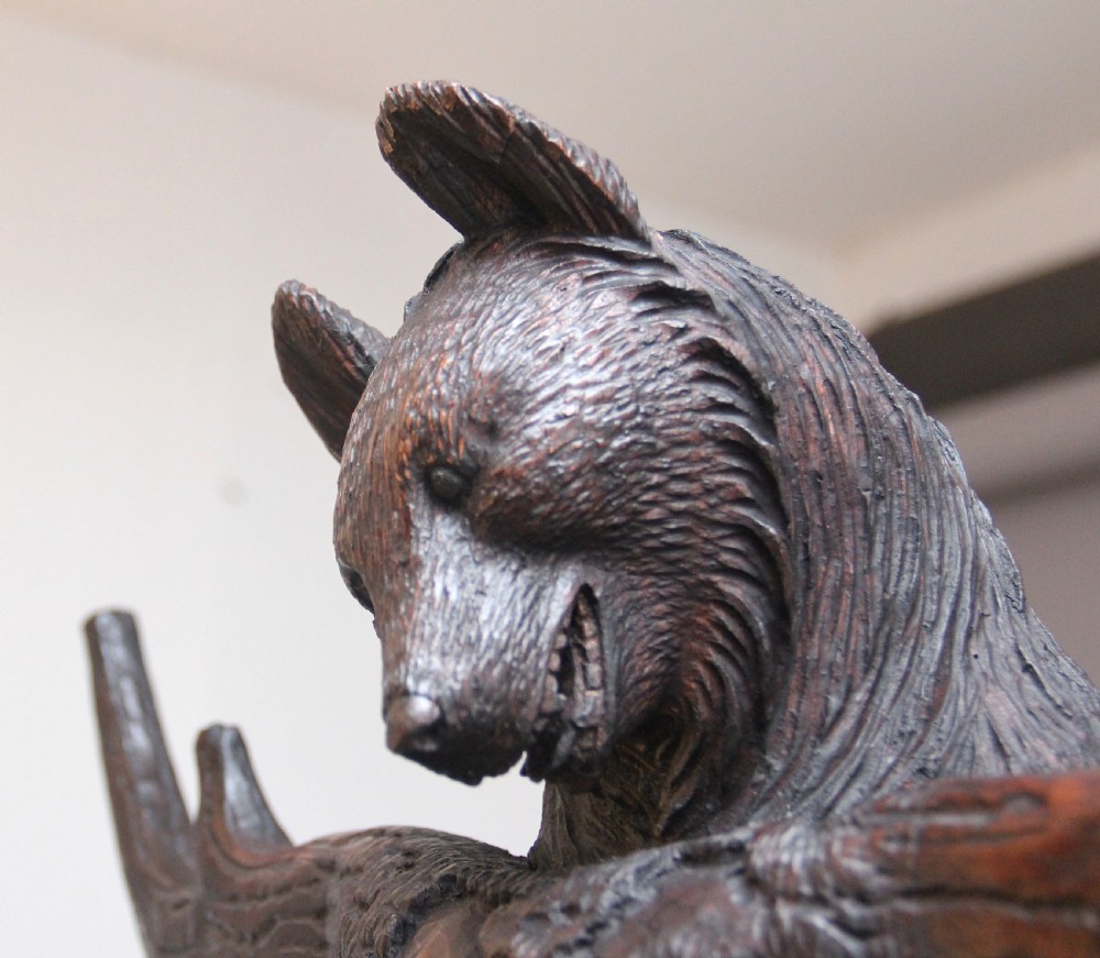 An early twentieth-century carved wooden Black Forest hall stand featuring a tree trunk stem with coat pegs, topped by a bear cub and supported at the base by a larger standing bear with an umbrella hoop and drip pan. View V