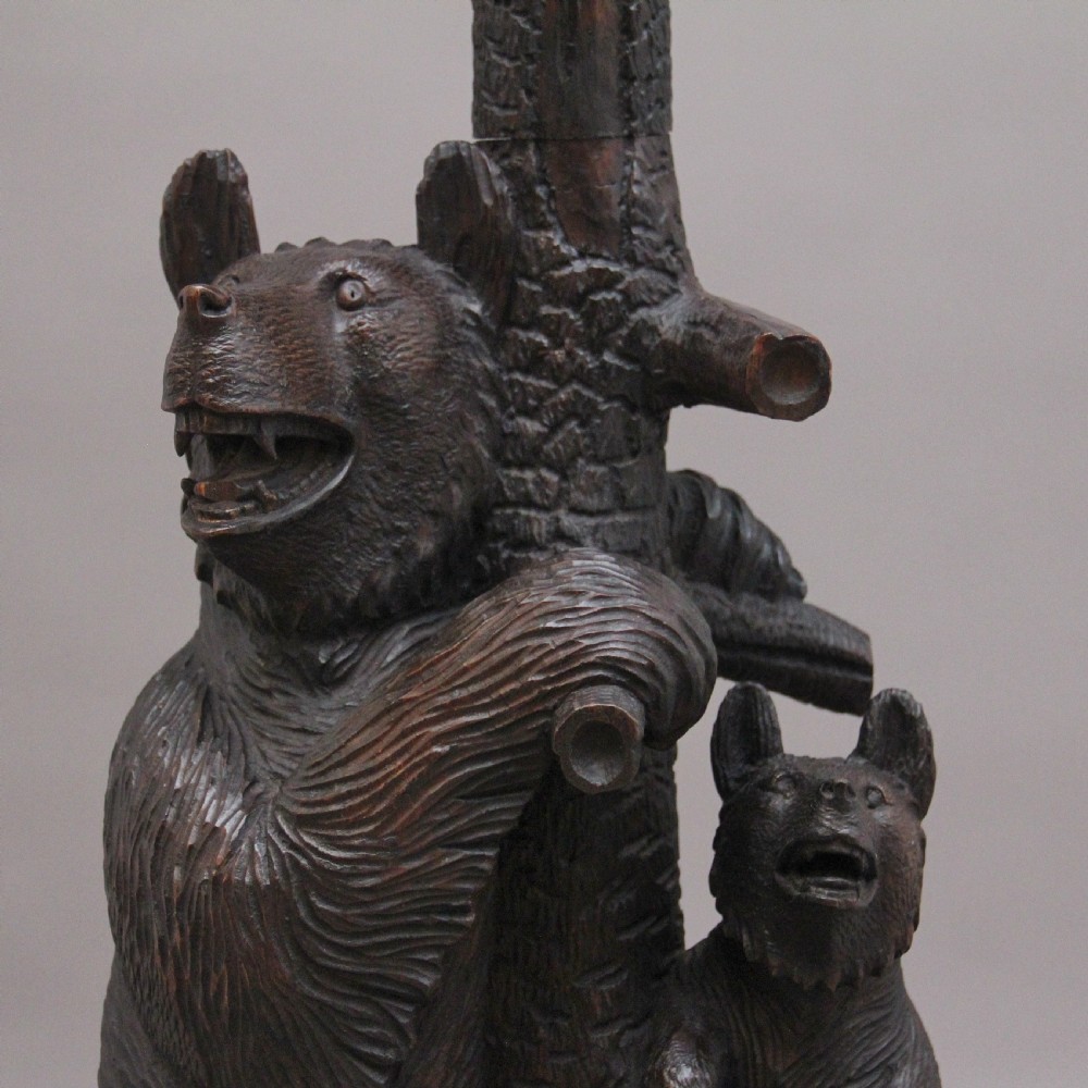 An early twentieth-century carved wooden Black Forest hall stand featuring a tree trunk stem with coat pegs, topped by a bear cub and supported at the base by a larger standing bear with an umbrella hoop and drip pan. View X