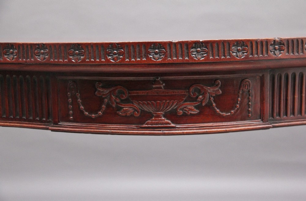 A dark mahogany serpentine console table with fluted legs and carved urn and garland motifs along the apron. View IX