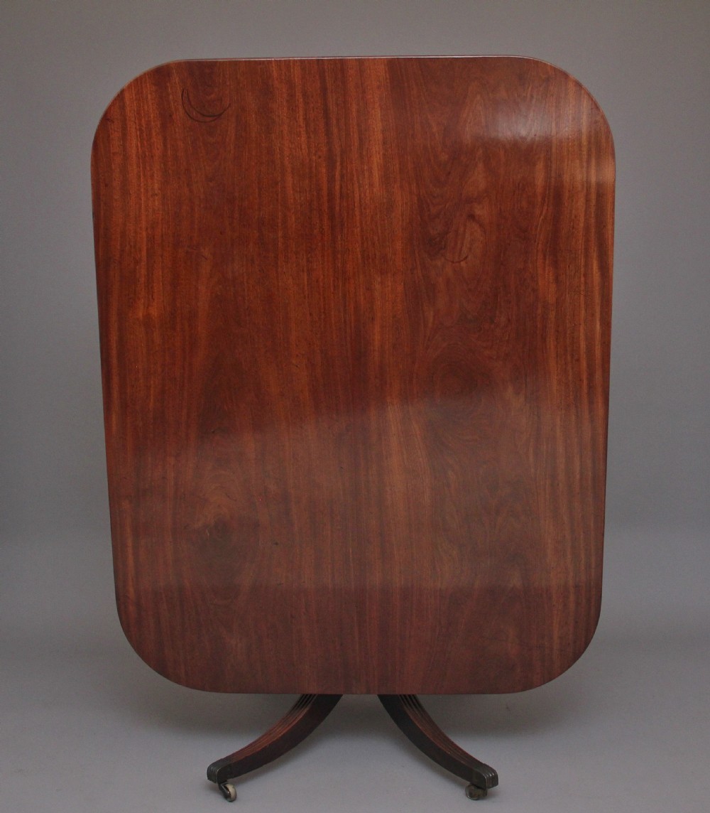 An early 19th-century mahogany tilt-top breakfast table with a rounded rectangular top shown in the upright position above a quadruped pedestal base with brass castors.
