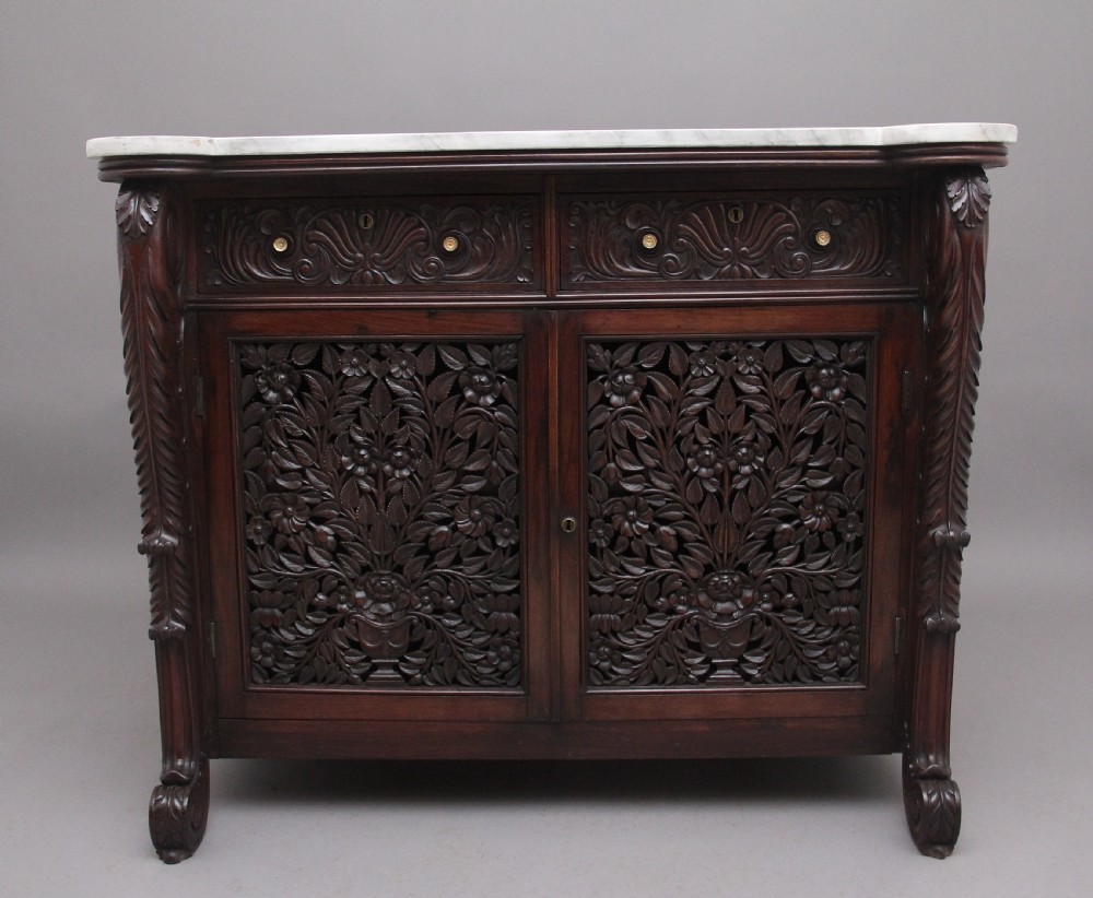 A 19th-century Anglo-Indian dark wood cabinet featuring a white marble top, two carved drawers, and two doors decorated with intricate pierced floral relief motifs.