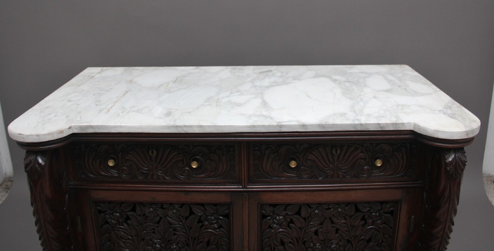 A 19th-century Anglo-Indian dark wood cabinet featuring a white marble top, two carved drawers, and two doors decorated with intricate pierced floral relief motifs. View X