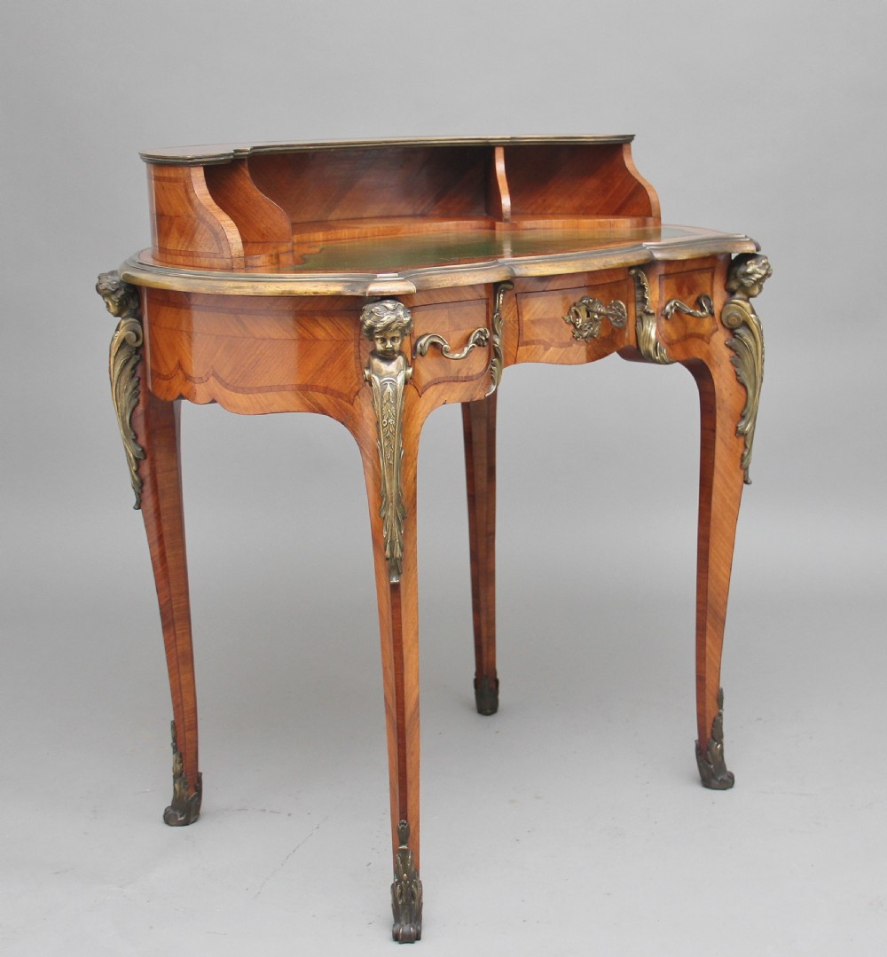 A 19th-century French kingwood writing table featuring ornate gilt-bronze mounts, cabriole legs, a green leather inset top, and a raised shelving superstructure.