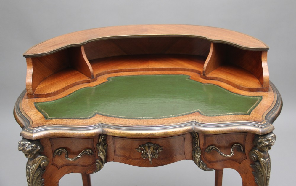 A 19th-century French kingwood writing table featuring ornate gilt-bronze mounts, cabriole legs, a green leather inset top, and a raised shelving superstructure. View VI