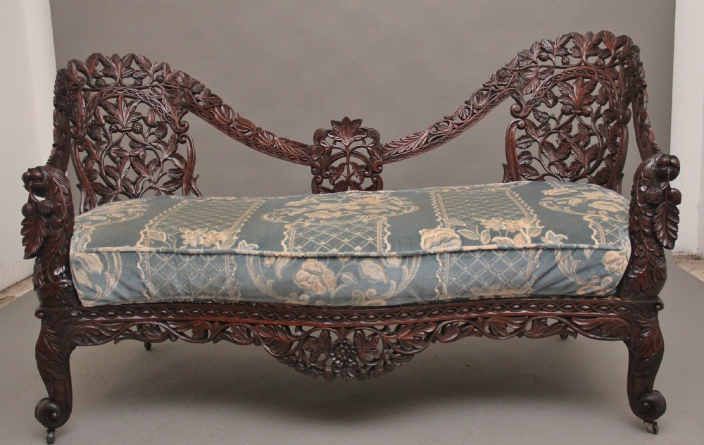 A 19th-century Anglo-Indian carved dark hardwood sofa featuring intricate foliage openwork, lion head armrests, and a patterned blue upholstered seat.