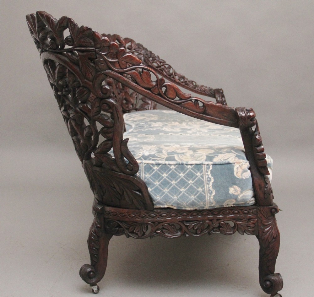A 19th-century Anglo-Indian carved dark hardwood sofa featuring intricate foliage openwork, lion head armrests, and a patterned blue upholstered seat. View XI