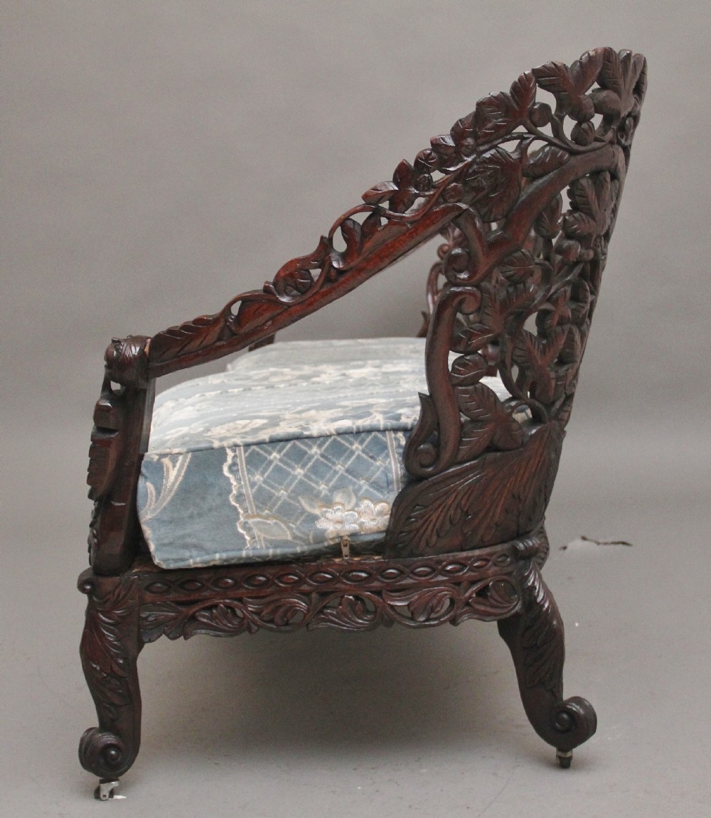 A 19th-century Anglo-Indian carved dark hardwood sofa featuring intricate foliage openwork, lion head armrests, and a patterned blue upholstered seat. View XIII