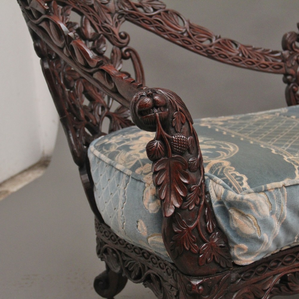 A 19th-century Anglo-Indian carved dark hardwood sofa featuring intricate foliage openwork, lion head armrests, and a patterned blue upholstered seat. View XIV