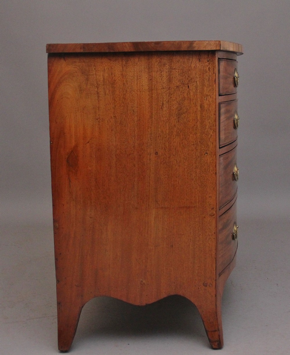 A nineteenth-century mahogany bowfront chest of drawers featuring four graduated drawers with oval brass handles and splayed feet. View VII