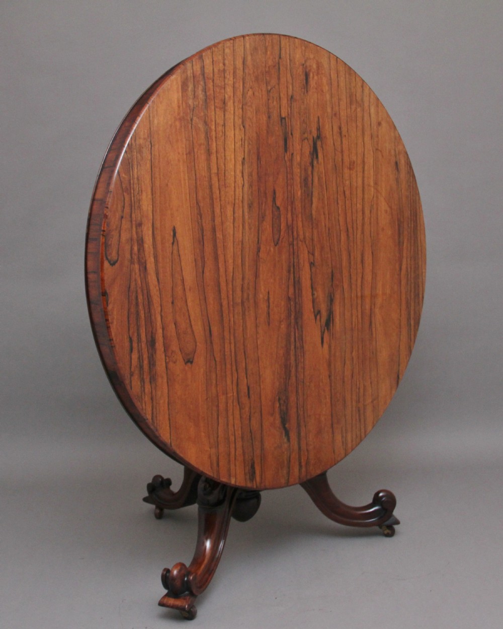 A 19th-century circular rosewood breakfast table displayed tilted upright on a carved tripod base with scroll feet.