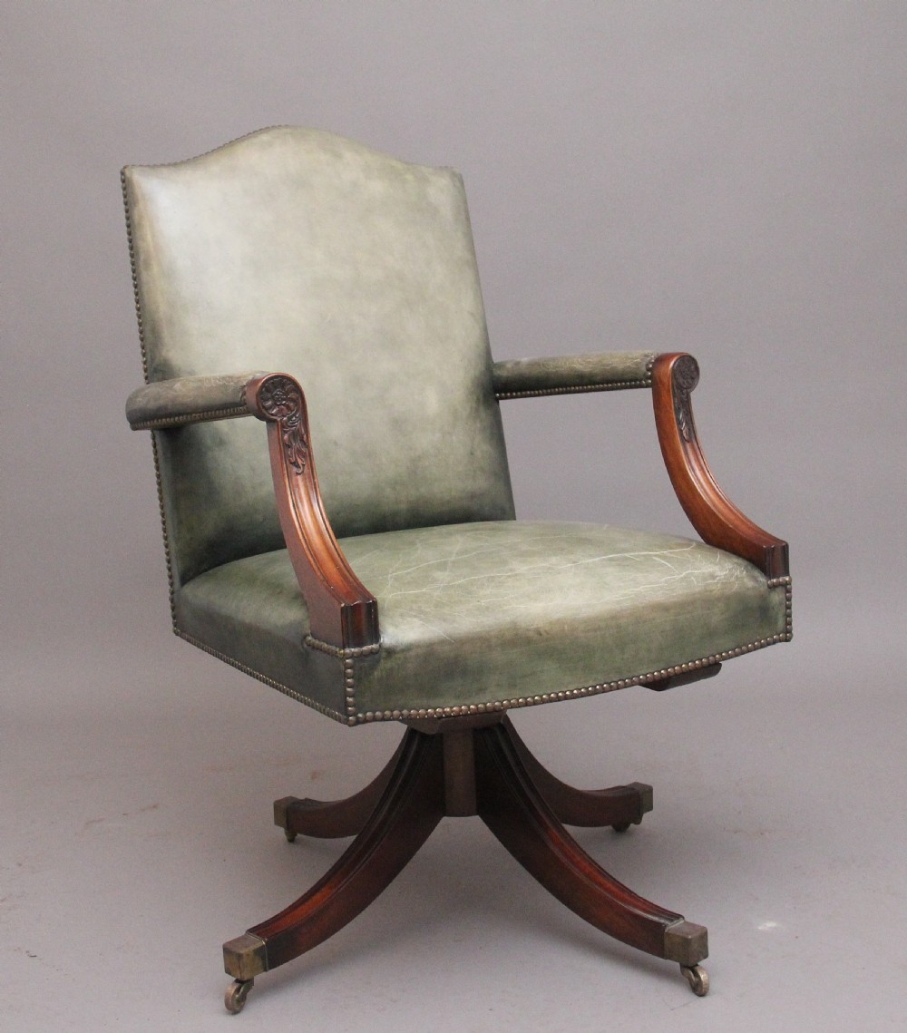 An early 20th century revolving desk chair featuring olive green leather upholstery with brass studding trim and carved mahogany armrests on a four-star base.