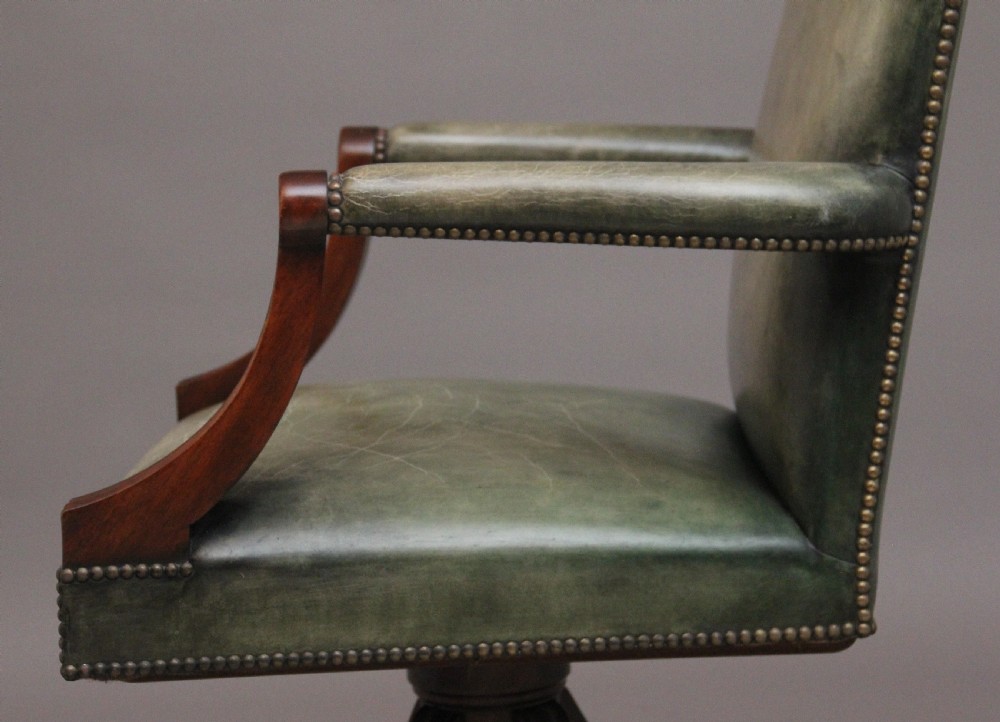 An early 20th century revolving desk chair featuring olive green leather upholstery with brass studding trim and carved mahogany armrests on a four-star base. View VII