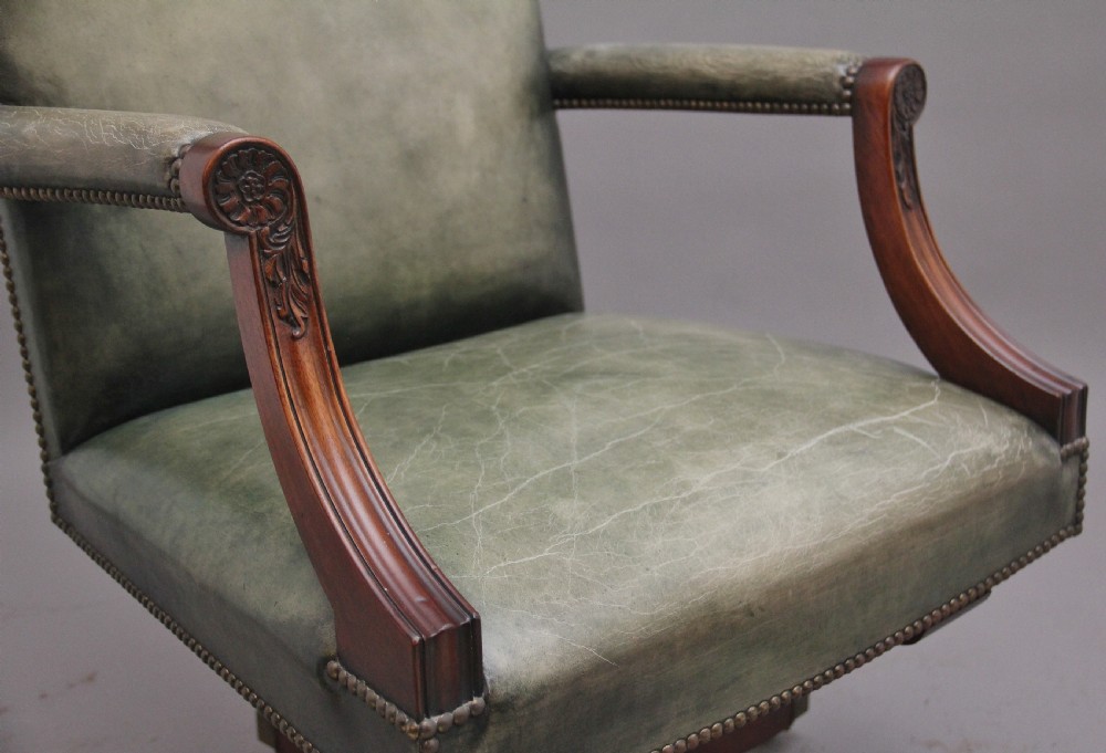 An early 20th century revolving desk chair featuring olive green leather upholstery with brass studding trim and carved mahogany armrests on a four-star base. View X