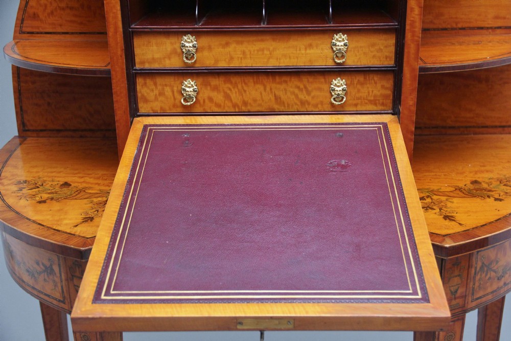 A 19th-century Sheraton revival satinwood writing desk featuring inlaid urn marquetry motifs, stepped side shelves, a central door compartment, and slender tapered legs. View VI