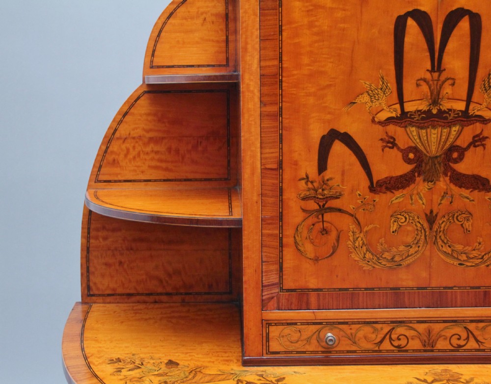 A 19th-century Sheraton revival satinwood writing desk featuring inlaid urn marquetry motifs, stepped side shelves, a central door compartment, and slender tapered legs. View X