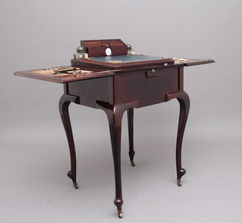 An early 20th-century dark mahogany metamorphic writing desk featuring cabriole legs on brass castors, fold-out side panels, a central leather writing slope, and an integrated clock with stationery compartment.