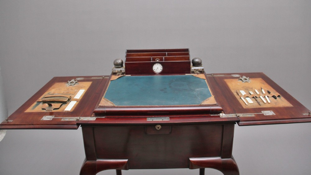 An early 20th-century dark mahogany metamorphic writing desk featuring cabriole legs on brass castors, fold-out side panels, a central leather writing slope, and an integrated clock with stationery compartment. View X