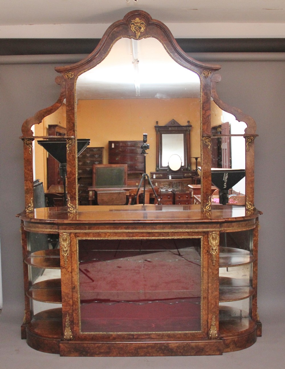 A 19th-century burr walnut credenza with an arched mirror back, ornate gilt-bronze mounts, open tier side shelving, and a central glazed compartment.