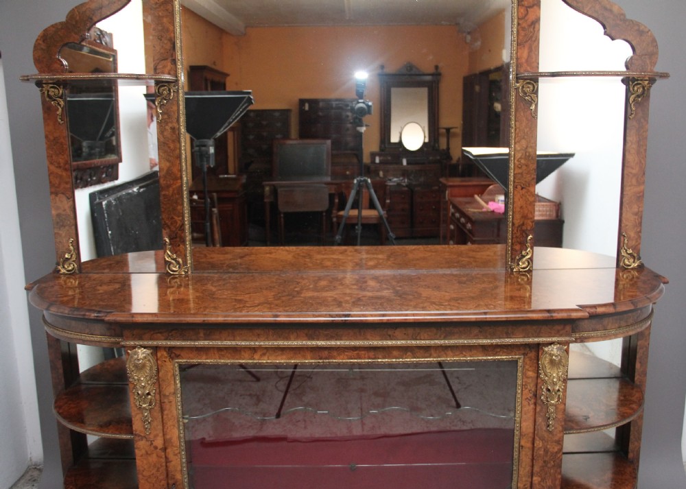 A 19th-century burr walnut credenza with an arched mirror back, ornate gilt-bronze mounts, open tier side shelving, and a central glazed compartment. View V
