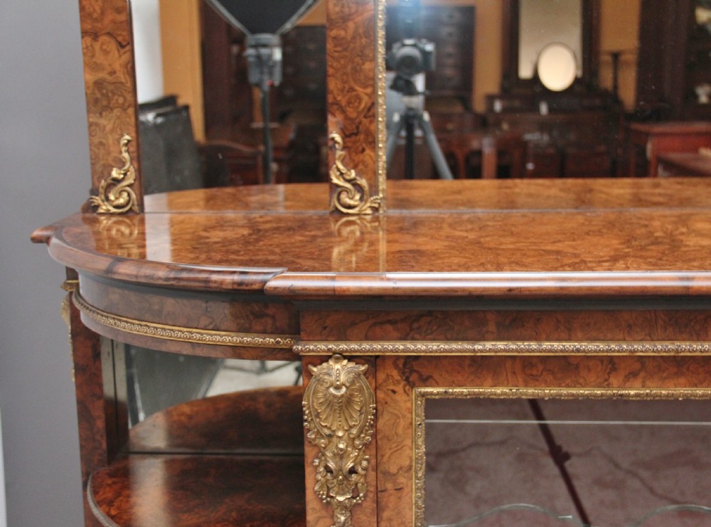 A 19th-century burr walnut credenza with an arched mirror back, ornate gilt-bronze mounts, open tier side shelving, and a central glazed compartment. View XII