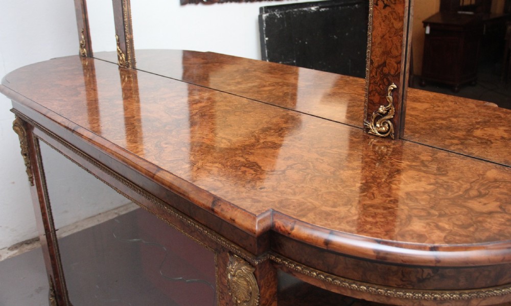 A 19th-century burr walnut credenza with an arched mirror back, ornate gilt-bronze mounts, open tier side shelving, and a central glazed compartment. View XIV