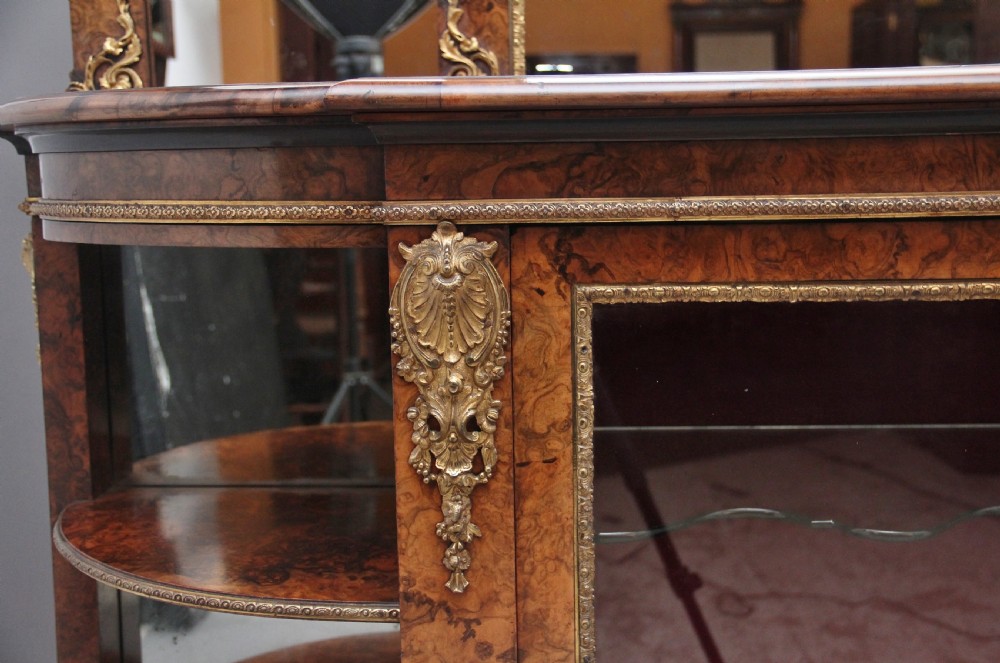 A 19th-century burr walnut credenza with an arched mirror back, ornate gilt-bronze mounts, open tier side shelving, and a central glazed compartment. View XIX