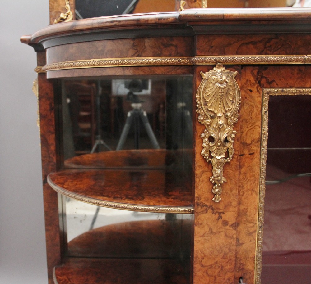 A 19th-century burr walnut credenza with an arched mirror back, ornate gilt-bronze mounts, open tier side shelving, and a central glazed compartment. View XX