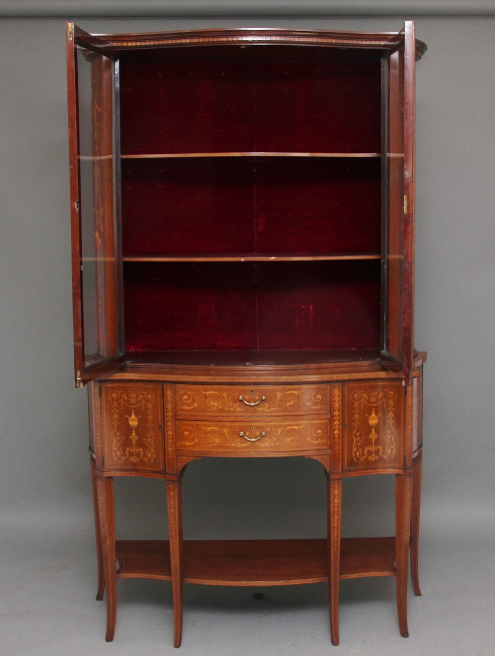 A late 19th-century bow-fronted mahogany display cabinet featuring glazed doors, satinwood marquetry inlay, slender square legs, and an undertier. View II
