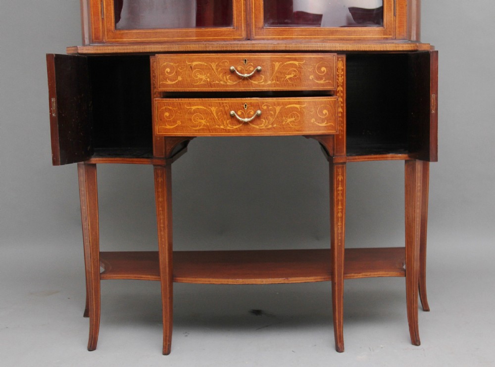 A late 19th-century bow-fronted mahogany display cabinet featuring glazed doors, satinwood marquetry inlay, slender square legs, and an undertier. View V