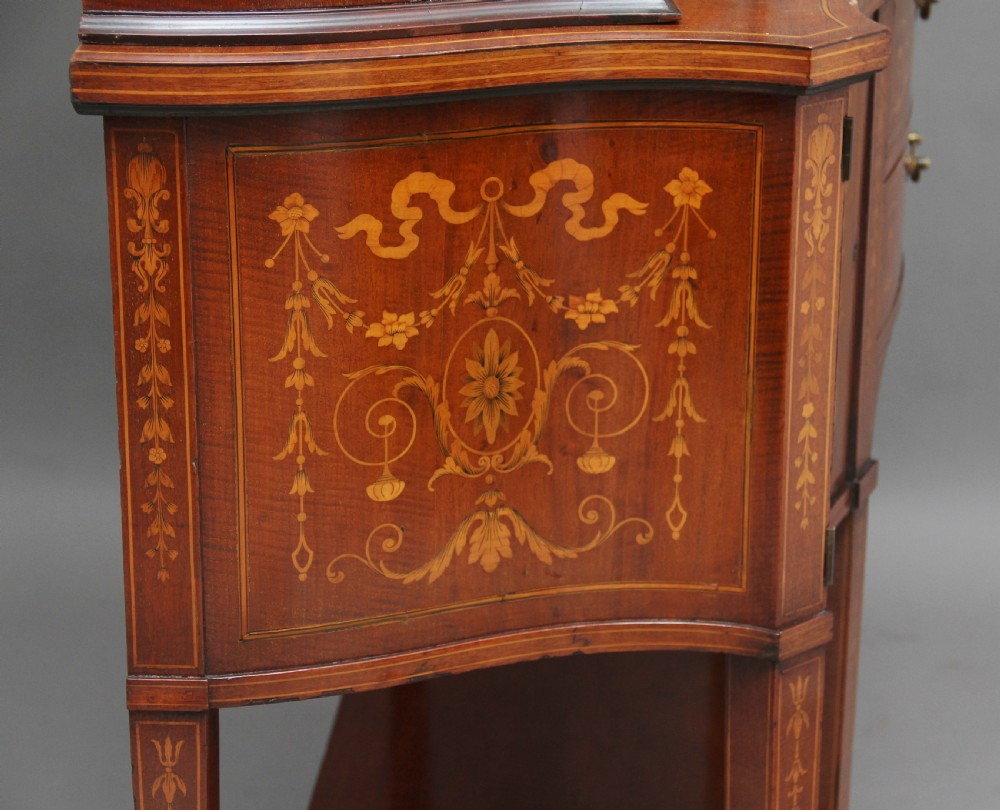 A late 19th-century bow-fronted mahogany display cabinet featuring glazed doors, satinwood marquetry inlay, slender square legs, and an undertier. View X