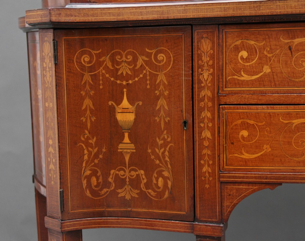 A late 19th-century bow-fronted mahogany display cabinet featuring glazed doors, satinwood marquetry inlay, slender square legs, and an undertier. View XII