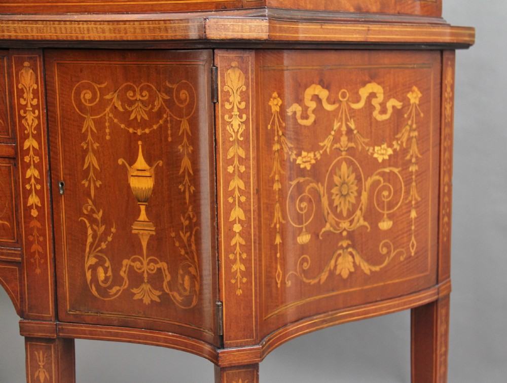 A late 19th-century bow-fronted mahogany display cabinet featuring glazed doors, satinwood marquetry inlay, slender square legs, and an undertier. View XIV