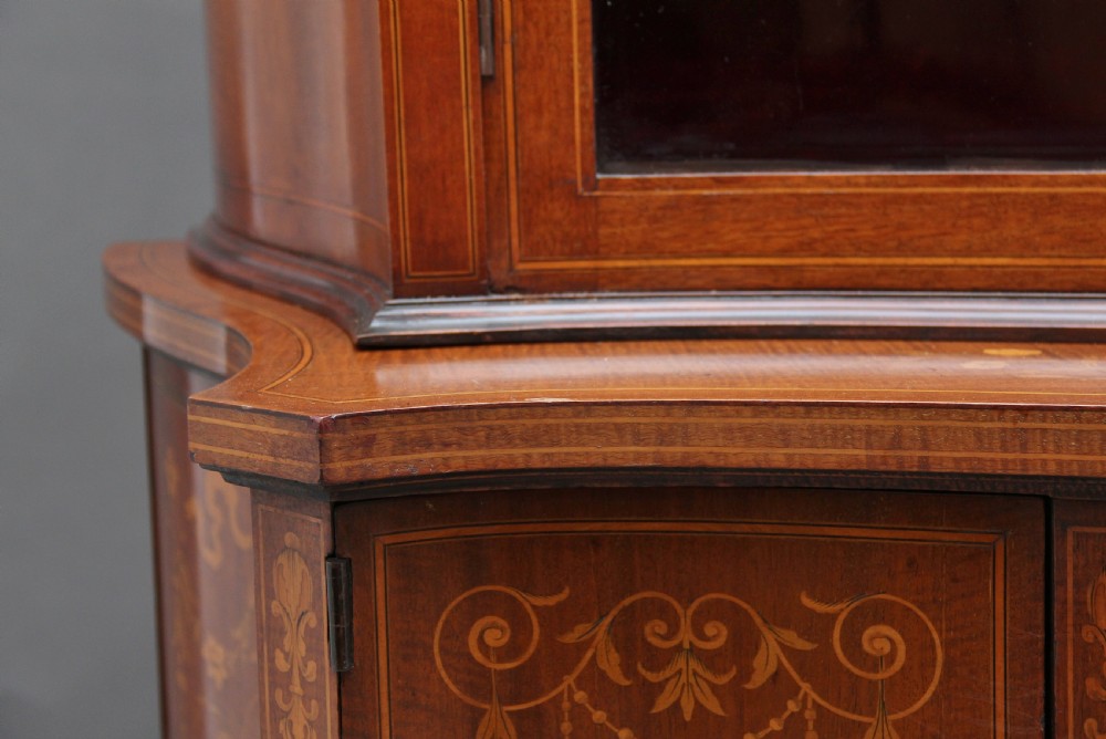 A late 19th-century bow-fronted mahogany display cabinet featuring glazed doors, satinwood marquetry inlay, slender square legs, and an undertier. View XV