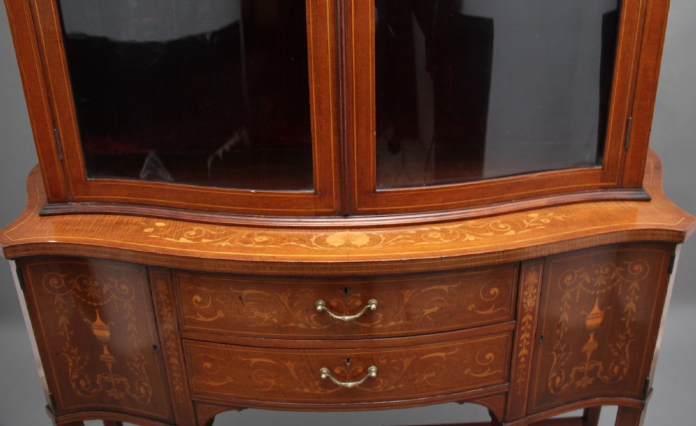 A late 19th-century bow-fronted mahogany display cabinet featuring glazed doors, satinwood marquetry inlay, slender square legs, and an undertier. View XVI