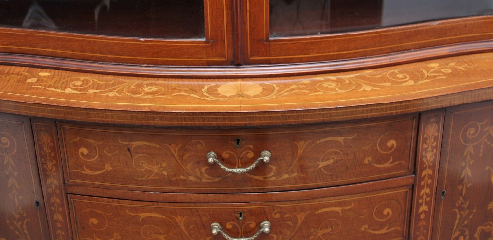A late 19th-century bow-fronted mahogany display cabinet featuring glazed doors, satinwood marquetry inlay, slender square legs, and an undertier. View XVII
