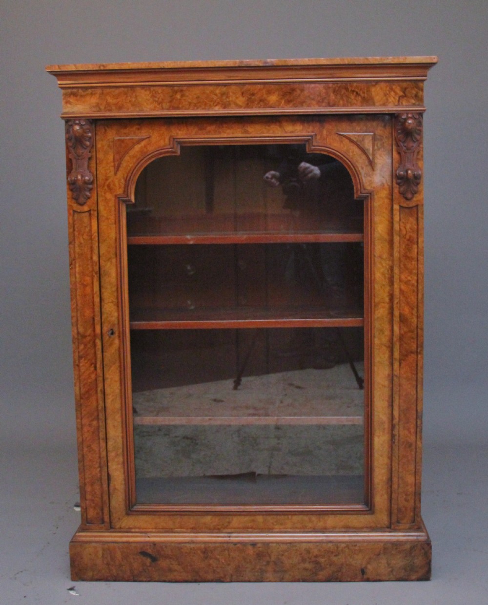 A Victorian burr walnut glazed cabinet featuring an arched glass door, carved corbels, three internal shelves, and a solid plinth base.