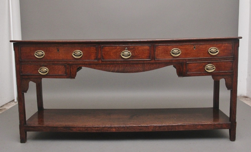An early 19th-century dark oak dresser base featuring five drawers with brass oval handles, an arched central apron, and a lower potboard shelf.