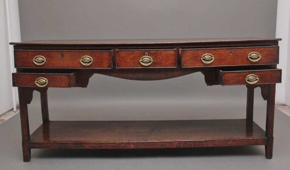 An early 19th-century dark oak dresser base featuring five drawers with brass oval handles, an arched central apron, and a lower potboard shelf. View II