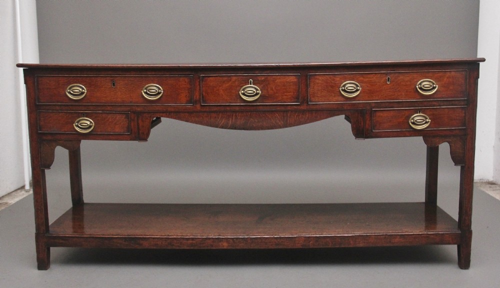 An early 19th-century dark oak dresser base featuring five drawers with brass oval handles, an arched central apron, and a lower potboard shelf. View VIII