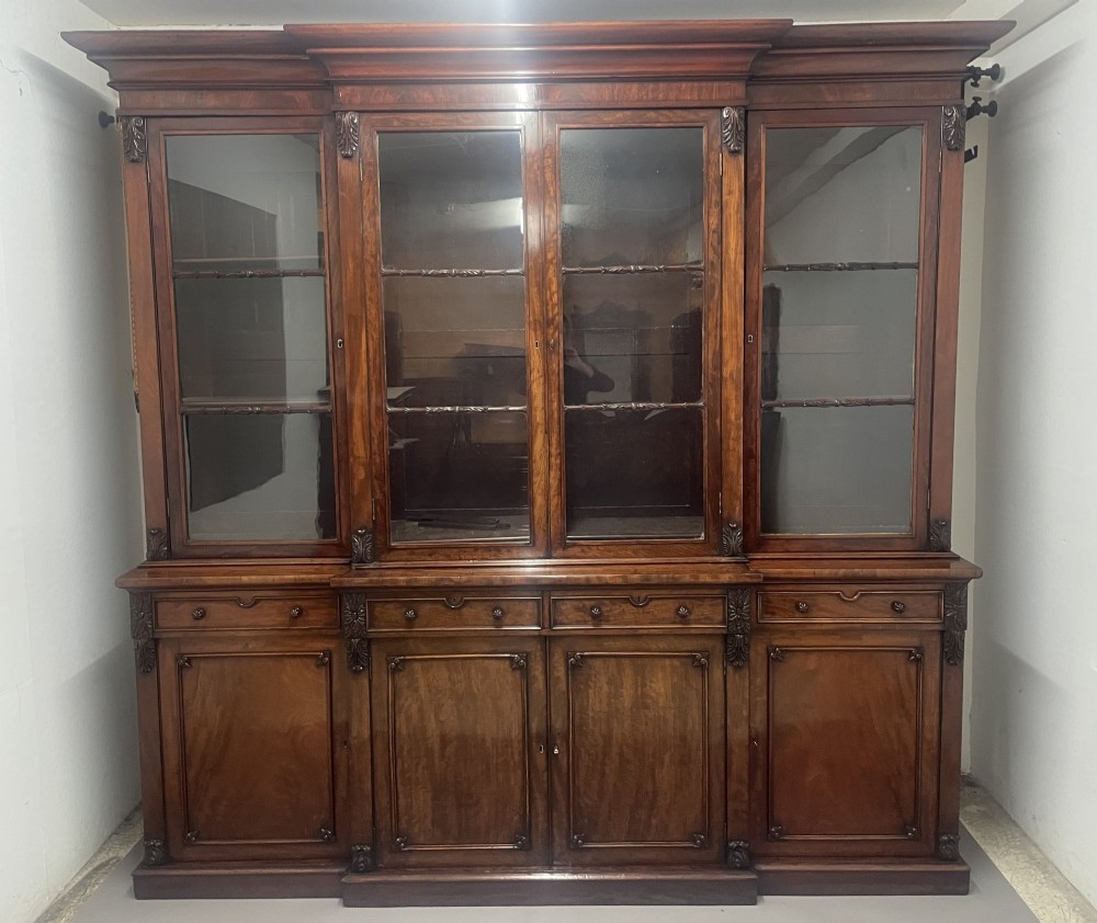 An early 19th-century dark mahogany breakfront bookcase featuring a four-door glazed upper section above four cupboard doors and drawers.