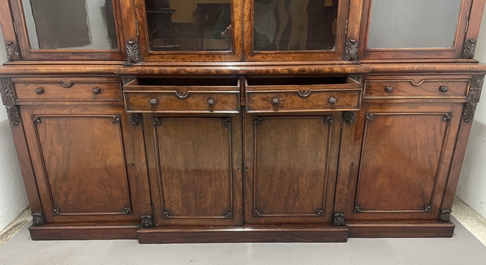 An early 19th-century dark mahogany breakfront bookcase featuring a four-door glazed upper section above four cupboard doors and drawers. View VII