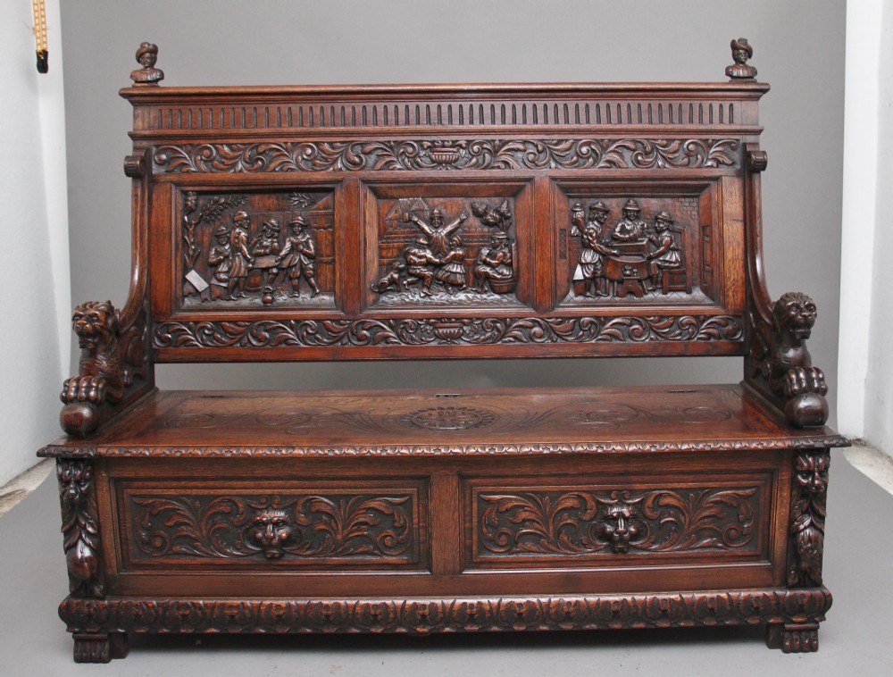 A 19th-century dark oak box settle with intricate relief-carved narrative panels, carved lion head armrests, and a hinged seat for internal storage.