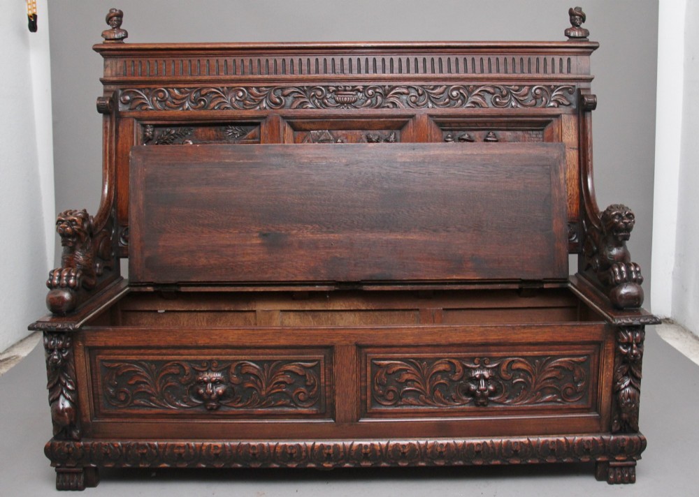 A 19th-century dark oak box settle with intricate relief-carved narrative panels, carved lion head armrests, and a hinged seat for internal storage. View II