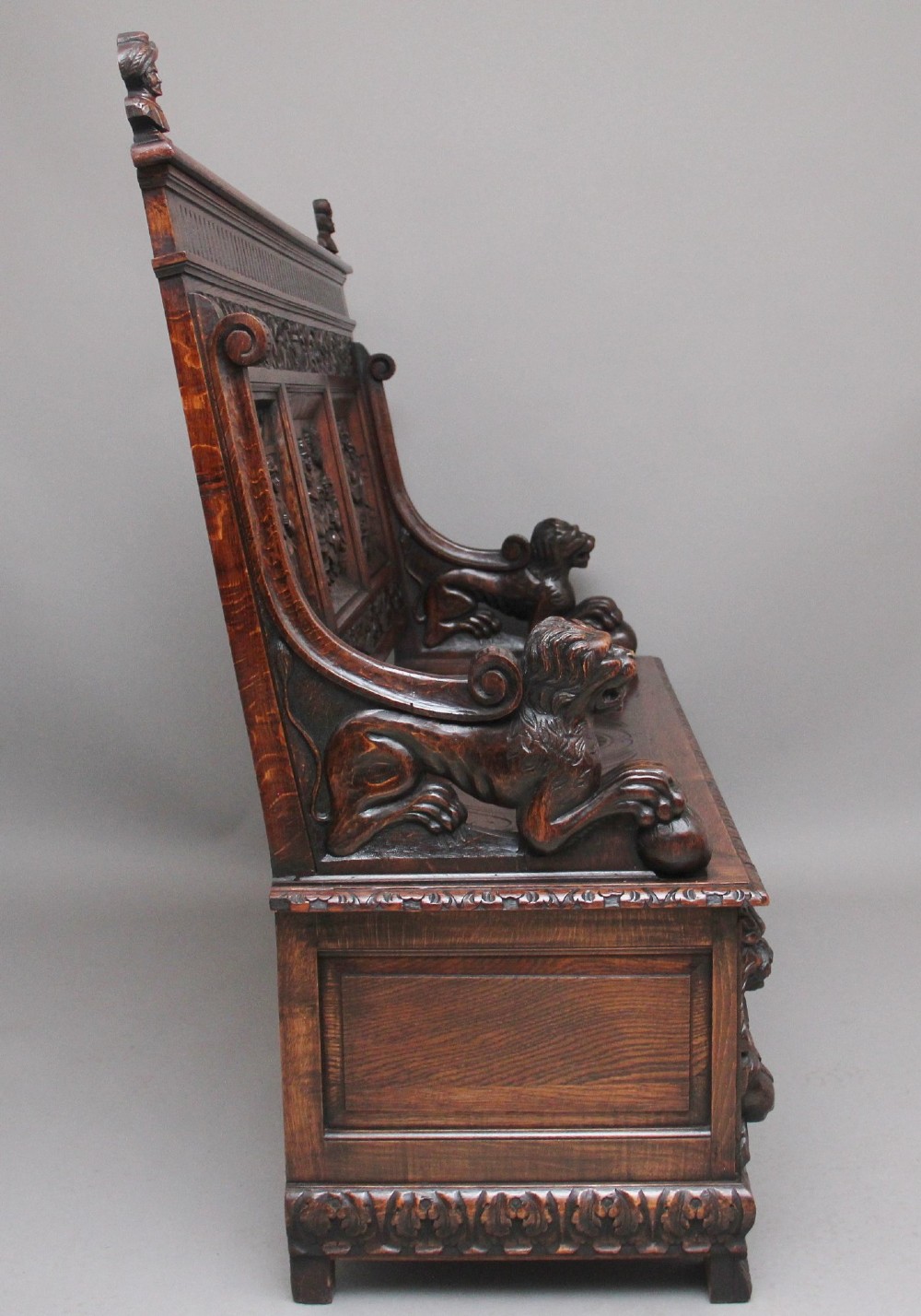 A 19th-century dark oak box settle with intricate relief-carved narrative panels, carved lion head armrests, and a hinged seat for internal storage. View III