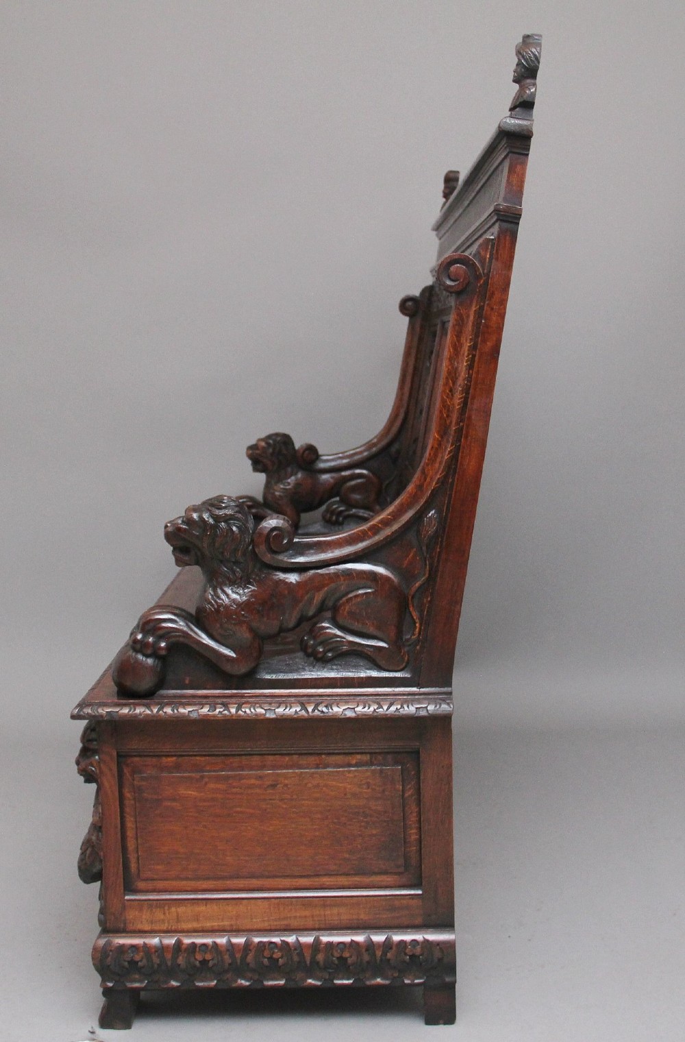 A 19th-century dark oak box settle with intricate relief-carved narrative panels, carved lion head armrests, and a hinged seat for internal storage. View V
