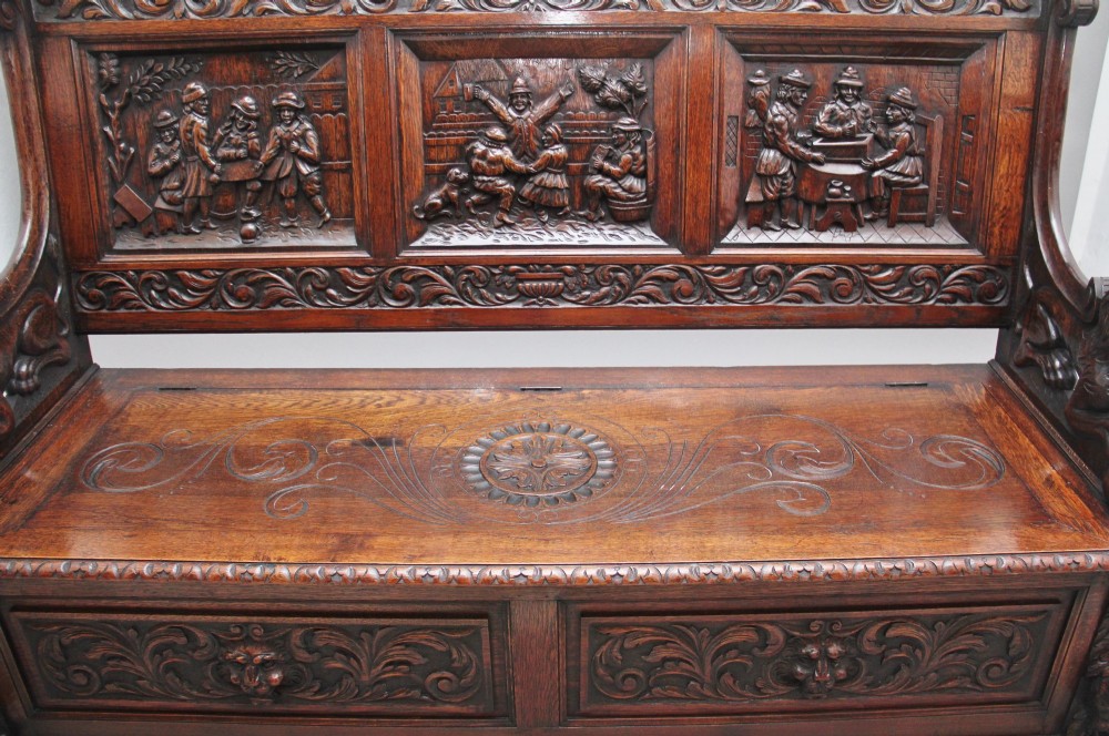 A 19th-century dark oak box settle with intricate relief-carved narrative panels, carved lion head armrests, and a hinged seat for internal storage. View VI