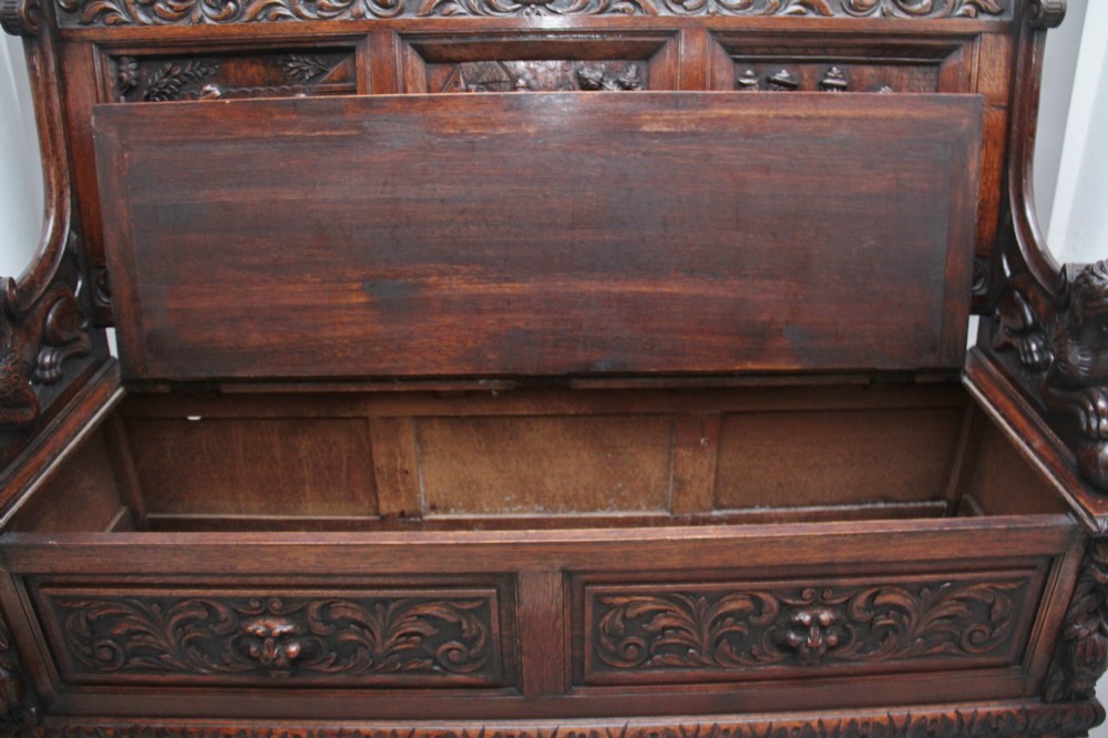 A 19th-century dark oak box settle with intricate relief-carved narrative panels, carved lion head armrests, and a hinged seat for internal storage. View VII